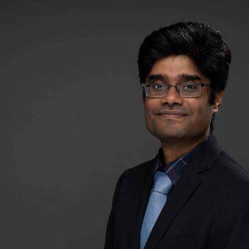 Rahat Rafiq in a black suit with a blue tie on a dark brown backdrop.